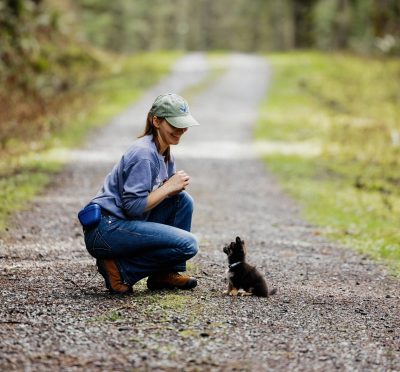 Straight on shot of Maleah, owner of Playful Balance Dog Training and Taquito, a long hair Chihuahua as a puppy on a gravel road.